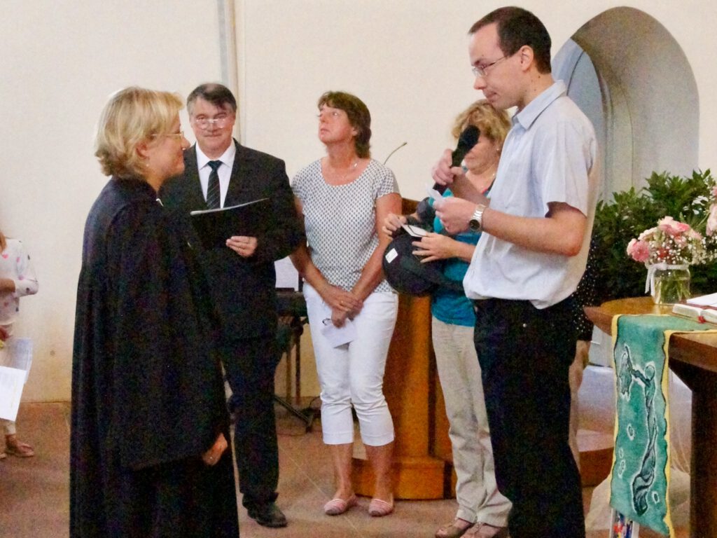 Eine Pfarrerin wird in der Evangelischen Martinskirche Neckarelz verabschiedet. Frank Heuß steht mit Mikrofon erhöht auf einem Podest und spricht in Richtung der Pfarrerin, die vor ihm steht. Im Hintergrund stehen drei Personen.