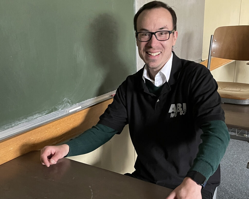 Frank Heuß sitzt in einem Klassenzimmer der Ludwig-Erhard-Schule Mosbach, an einem Einzeltisch. Er trägt das Abschluss-Shirt des Abiturjahrgangs 2023 der Schule über der Alltagskleidung.