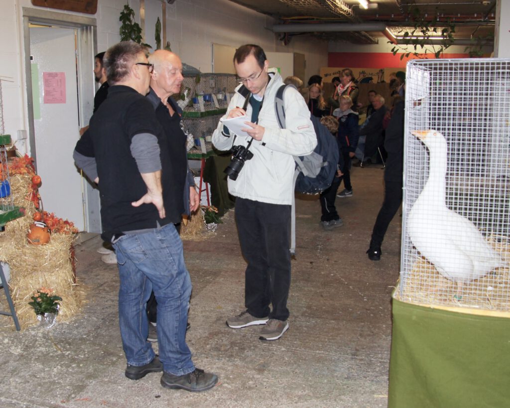 Der Journalist Frank Heuß (vorne 3.v.r.) im Gespräch mit zwei Personen in schwarzen Vereins-Shirts. Er schreibt in einem Notizblock, trägt eine Kamera um den Hals und einen Rucksack über die Schulter. Rechts ist eine Ganz in einem Käfig zu sehen, im Hintergrund Besucherinnen und Besucher der Kleintierschau (Archivaufnahme). Foto: pr
